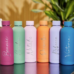 Könnte beinhalten: Sechs farbige Wasserflaschen in Pink, Grün, Hellrosa, Orange, Lavendel und Blau. Jede Flasche hat eine silberne Namensaufschrift: Bonnie, Caroline, Natalie, Priscella, Rebecca und Victoria. Die Flaschen stehen auf einer dunklen Holzoberfläche.