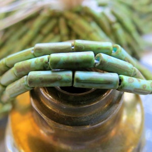 May include: Close-up of a necklace made of cylindrical beads. The beads are primarily green with brown and blue accents. The necklace is strung together, and the beads are arranged in a curved shape. The background is blurred, with more green beads visible.