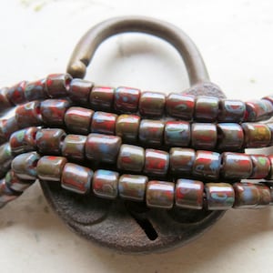 May include: A close-up of a vintage padlock wrapped with multiple strands of brown, red, and blue glass beads. The beads have a speckled, iridescent finish.