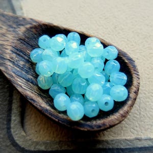 May include: A close-up of a wooden spoon filled with light blue iridescent glass beads. The beads are faceted and have a slightly opaque finish.