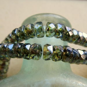 May include: Two strands of faceted rondelle beads featuring a metallic, iridescent finish with deep olive green and bronze tones. The beads are strung on a clear filament, displaying a reflective, multi-faceted surface that catches the light.