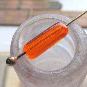 May include: An orange glass bead with a cylindrical shape is pierced by a thin metal wire. The bead is suspended above the opening of a vintage, light purple glass bottle. The bead's color is a vibrant, translucent orange.