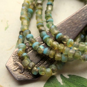 May include: Close-up of beaded necklaces in shades of green, blue, and gold. The beads are strung together and rest on a vintage silver spoon. The beads have a faceted texture, and the colors create an iridescent effect.