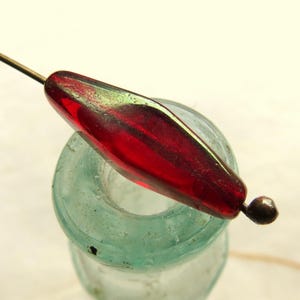 May include: A long, tapered, translucent red glass bead with an iridescent green sheen, threaded onto a thin metal pin. The bead has a smooth, polished surface and a pointed shape, featuring a small dark metal stopper at the end of the pin.