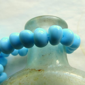 Könnte beinhalten: Eine Schnur aus hellblauen Glasperlen, möglicherweise für die Schmuckherstellung, auf einer strukturierten Glasflasche.