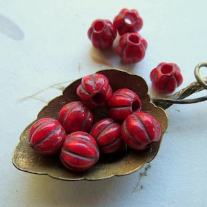 Puede incluir: Una colección de cuentas de vidrio rojo con textura acanalada, expuestas en una cuchara de hoja dorada. Las cuentas tienen un diámetro aproximado de 1 cm.