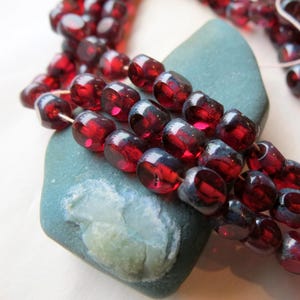 May include: A strand of dark red glass beads with a slightly iridescent finish. The beads are round and have a slightly flattened shape. The beads are strung on a wire and are lying on a gray rock.