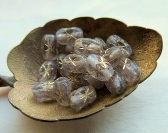 May include: A collection of small, oval-shaped, translucent light purple beads with etched gold-coloured star patterns, resting inside a decorative metal spoon shaped like a leaf with a textured, matte gold-toned finish and scalloped edges.