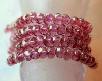 May include: Close-up of a strand of faceted, pink beads. The beads are strung together, forming multiple loops. The beads have a sparkling, reflective quality, and the colour is a vibrant, rosy pink. The beads are resting on a white surface.