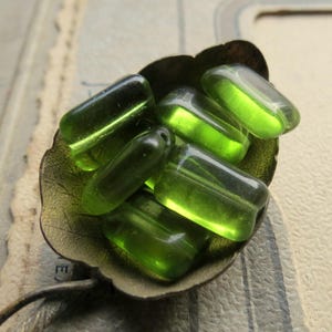 Könnte beinhalten: Nahaufnahme einer Sammlung transluzenter, olivgrüner Glasperlen. Die Perlen sind rechteckig mit abgerundeten Kanten und befinden sich in einer dunklen, blattförmigen Schale. Die Perlen haben eine glänzende Oberfläche.