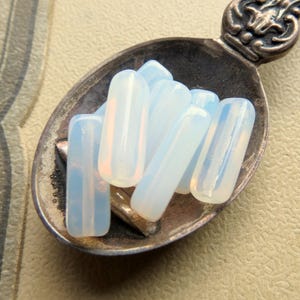 May include: A selection of opalescent, rectangular beads with rounded edges, presented in a vintage silver spoon. The beads exhibit a soft, translucent quality with delicate pink and blue tones, possibly made of gemstone or glass.