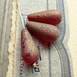 May include: Three red teardrop-shaped beads with a gold speckled finish. The beads are strung on a thin gold wire, resting on a vintage paper background. The beads have a translucent quality, and a small gold bead is at the base of each.