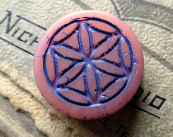 May include: A round, pink bead with a blue floral design etched into its surface. The bead has a slightly cracked texture and a raised design. The background is a textured, tan surface with faded text.