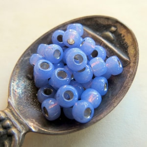 May include: A collection of small, round, light blue beads with a translucent, iridescent finish and a silver-toned core. The beads are piled together inside the bowl of a tarnished, antique-style metal spoon with a textured handle.
