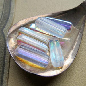May include: A collection of iridescent glass beads in a silver spoon. The beads are oblong, with a clear, textured surface that reflects a rainbow of colors. The spoon is made of silver and has a simple, elegant design. The beads are a craft supply.