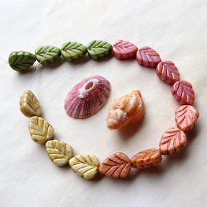 May include: A strand of leaf-shaped glass beads in shades of green, pink, and orange. The beads are strung on a clear thread and are arranged in a circle. There are also two seashells in the image, one pink and one orange.