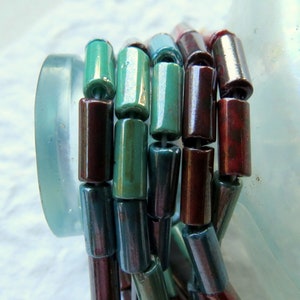 May include: Close-up of a stack of iridescent glass beads. The beads are cylindrical and have a metallic sheen. The colors of the beads are teal, turquoise, and burgundy.