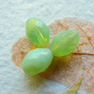 May include: Three green glass beads with a faceted finish. The beads are oval shaped and have a translucent appearance.