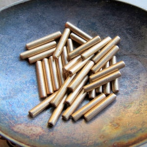 May include: A collection of small, cylindrical gold-colored beads. The beads are scattered in a silver-toned bowl. The beads have a matte finish and are evenly sized. These beads are suitable for jewelry making and crafting projects.