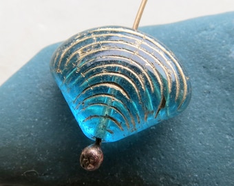 May include: A blue glass bead with gold accents, shaped like a seashell. The bead has a hole in the top for stringing.