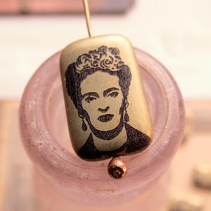 Puede incluir: Un colgante rectangular dorado con un retrato negro de Frida Kahlo. El colgante está suspendido por un fino alambre dorado y una pequeña cuenta de color cobre. El colgante se encuentra sobre un fondo rosa suave.