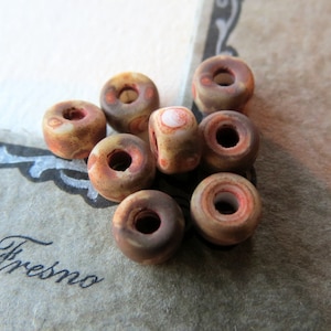 May include: A group of eight brown and white ceramic donut beads with red and white accents. The beads are arranged in a pile on a brown surface.