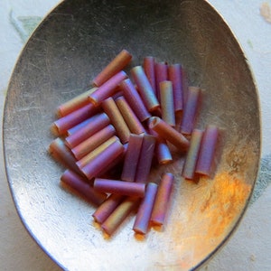 May include: A pile of iridescent glass beads in shades of purple and brown. The beads are cylindrical and have a smooth, glossy finish.