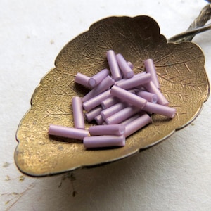 May include: A pile of small, light purple glass beads in the shape of cylinders. The beads are in a small, gold-colored dish.