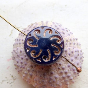 Puede incluir: Un botón redondo azul oscuro con un intrincado diseño en espiral en un color más claro. Un fino alambre de color cobre atraviesa el botón. El botón descansa sobre una superficie texturizada de color púrpura claro.