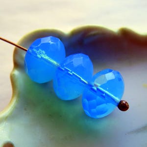 May include: Close-up of three faceted, oval-shaped, bright blue beads strung on a thin wire. The beads have a translucent quality and are arranged in a row. The background is a blurred, light green and white surface.
