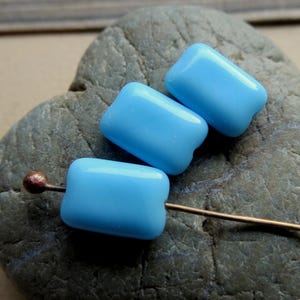 May include: Three smooth, rectangular, light blue beads with rounded corners and a glossy finish. One bead is threaded onto a thin, metallic pin with a small, round, copper-colored head, while the other two beads are positioned directly above it.