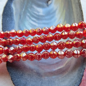 Könnte beinhalten: Eine Strähne roter Glasperlen mit einem metallischen Schimmer. Die Perlen sind facettiert und haben eine leicht unregelmäßige Form. Die Perlen sind auf einem durchsichtigen Faden aufgefädelt und werden auf einem weißen Hintergrund präsentiert.