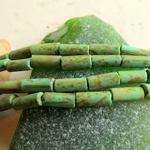 May include: A strand of green glass beads with a mottled, aged finish. The beads are cylindrical in shape and have a rustic, earthy look.