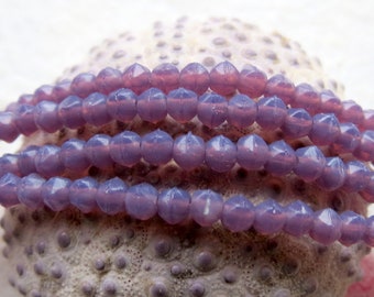 May include: A strand of light purple glass beads with a faceted, star-shaped design. The beads are arranged in a circular pattern on a textured surface.