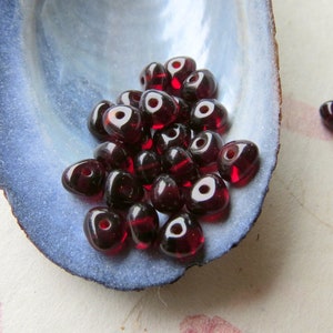 May include: A pile of small, dark red glass beads in the shape of a teardrop. The beads have a hole in the center and are arranged in a pile on a blue and white seashell.