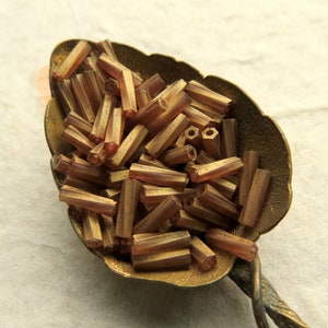 May include: A close-up of a spoon filled with small, brown, glass beads. The beads are hexagonal in shape and have a metallic sheen.