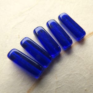May include: Five cobalt blue glass beads, each with a rounded rectangular shape. The beads are arranged diagonally, showcasing their glossy, transparent quality. These beads are suitable for jewelry making and crafting.