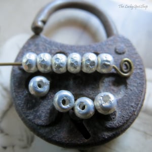 May include: A close-up of silver metal beads with a hammered texture. The beads are arranged in a line on a rusty metal surface. There are also several loose beads on the surface.
