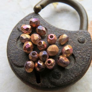 May include: A collection of small, iridescent glass beads in shades of copper and pink. The beads are faceted and have a metallic sheen. They are arranged on a rusty metal surface.