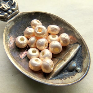 Puede incluir: Una colección de cuentas de perlas de color rosa pálido en una cuchara de plata vintage. Las cuentas son redondas y tienen un acabado liso y brillante.