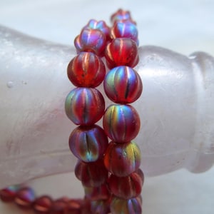 Puede incluir: Una hebra de cuentas de vidrio rojo iridiscente con textura acanalada. Las cuentas son redondas y tienen un brillo metálico.