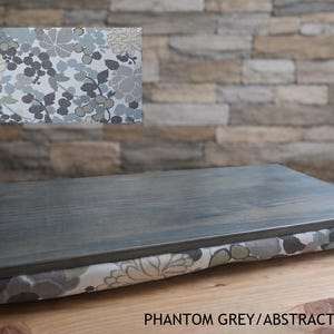May include: A gray wooden lap desk with a floral patterned cushion. The top is a solid gray, and the cushion features a pattern of gray, white, and green flowers. The text "Phantom Grey/Abstract Flowers" is visible.