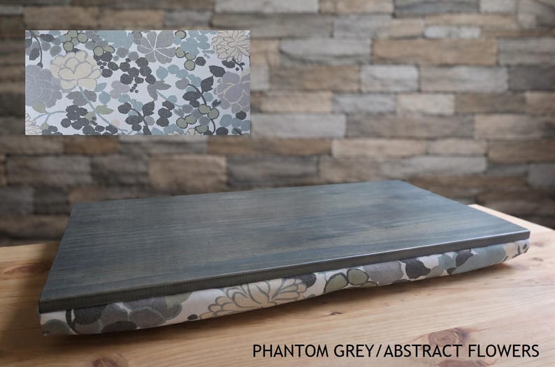 May include: A gray wooden lap desk with a floral patterned cushion. The top is a solid gray, and the cushion features a pattern of gray, white, and green flowers. The text "Phantom Grey/Abstract Flowers" is visible.