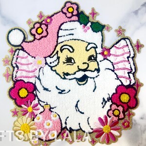 Puede incluir: Parche bordado de hierro rosa y blanco de un Papá Noel sonriente con un gorro rosa y una barba blanca. El parche está rodeado de flores rosas y amarillas y purpurina dorada.