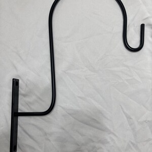 May include: Black metal wall-mounted hook with a curved arm and a small hook at the end. The hook is designed to hold a variety of items, such as plants, lanterns, or other decorative items.