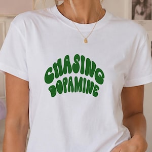 May include: A white short-sleeved t-shirt featuring the text "CHASING DOPAMINE" printed in a dark green, wavy, retro-style font across the chest. The shirt has a classic crew neckline and a straight hem, paired with light blue denim jeans.