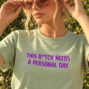 以下が含まれることがあります： 胸元に太字のサンセリフ体で紫色の「THIS B*TCH NEEDS A PERSONAL DAY」というテキストがプリントされた、ライトセージグリーンの半袖Tシャツ。クラシックなクルーネックで、標準的でリラックスしたフィット感の構造をしています。