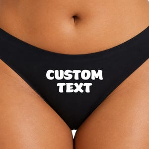 以下が含まれることがあります： 中央に太字の白いフォントで「CUSTOM TEXT」という文字がプリントされた黒い下着。この衣類は滑らかで無地の黒い生地で作られており、クラシックなカットが施されているため、シンプルで清潔感のある外観に仕上がっています。