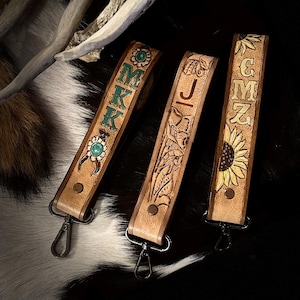 May include: Three leather keychains with personalized designs. One features the letters "MKK" in green, another has the letter "J" with floral details, and the last displays "GMZ" with a sunflower. Each keychain has a metal clasp.