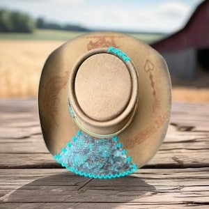 May include: A brown cowboy hat with turquoise and white trim. The hat has a design of a horse and rider on the crown.